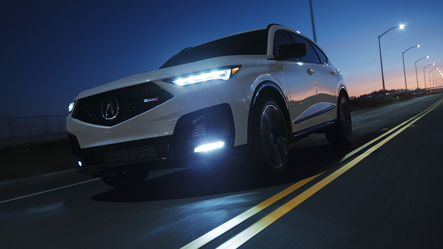 A white 2025 MDX Type S drives on an illuminated city highway at night with headlights on.