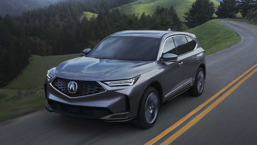 3/4 front view of grey MDX driving on a winding countryside highway with rolling hills and coniferous trees in the background. 