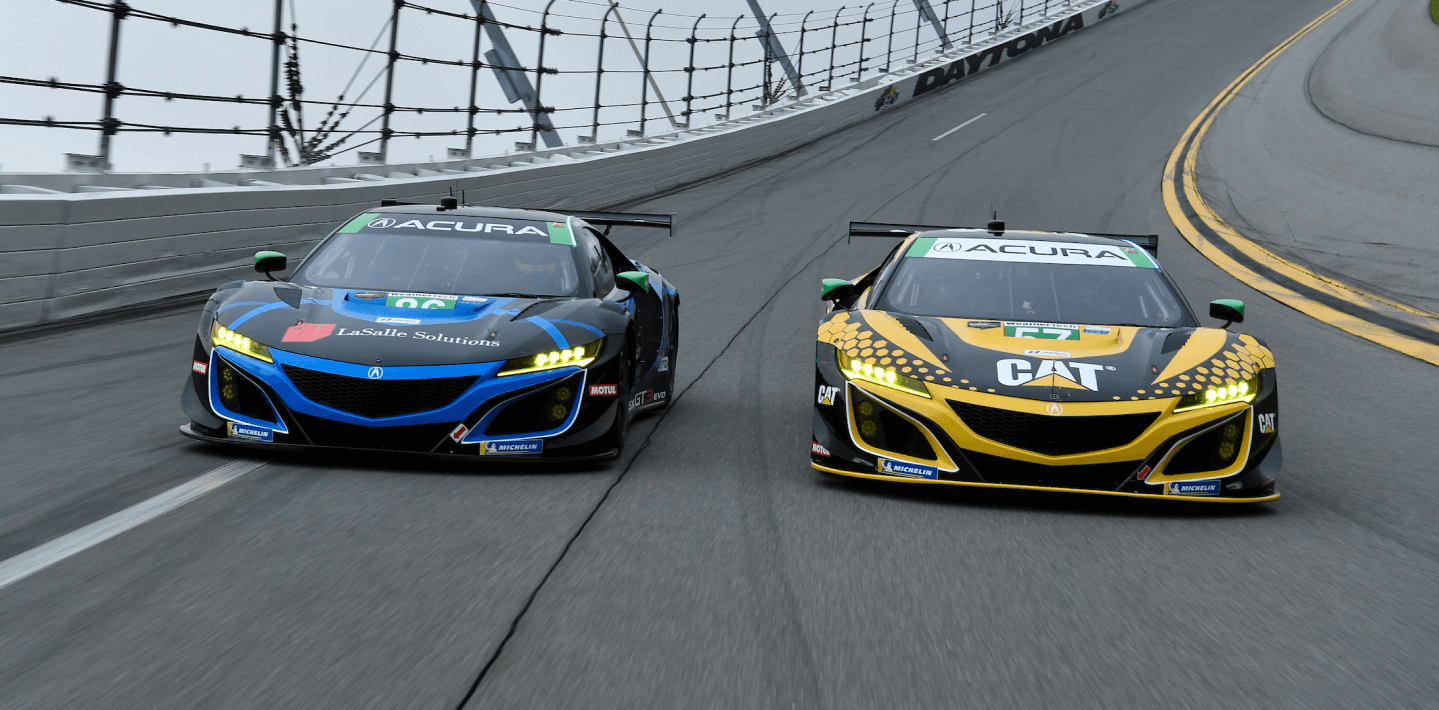 Front view of two Acura NSX GT3s, one blue and one red, both covered in sponsorship wraps, racing head-to-head on a track.  	Vue avant de deux Acura NSX GT3, l’une bleue et l’autre rouge, recouvertes de logos de commanditaires coursant sur un circuit de course.